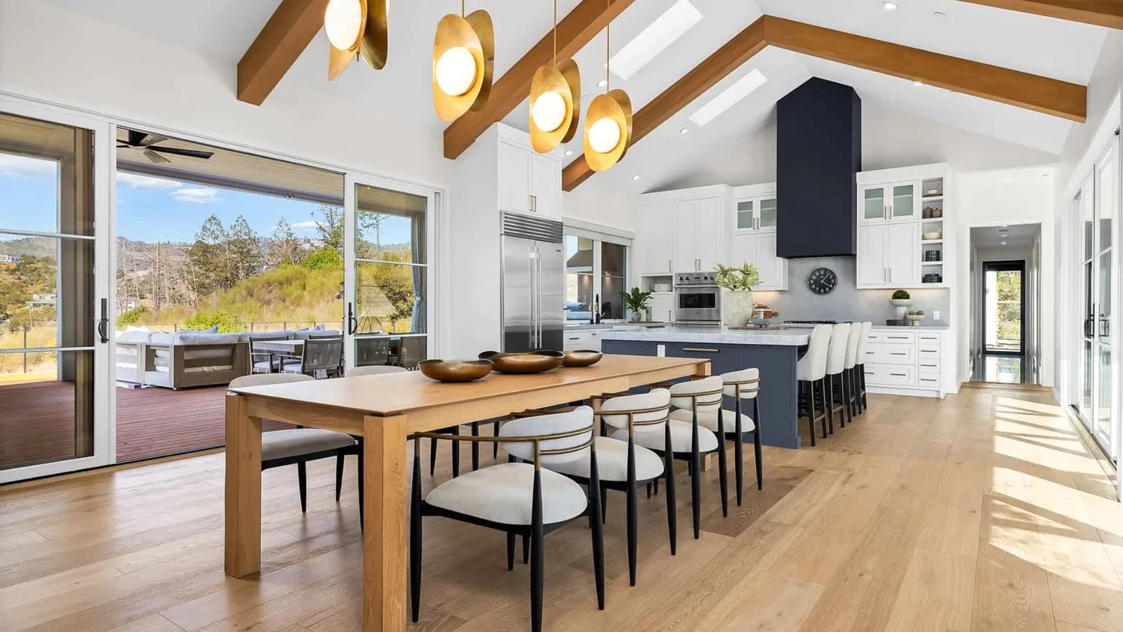 Modern open-plan outdoor kitchen and lounge with blue cabinetry, built-in grill, dining benches, and seating area under vaulted ceiling with skylights