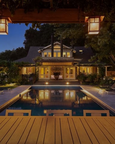 Lit craftsman house at dusk reflected in a calm pool, framed by pergola lanterns and mature trees
