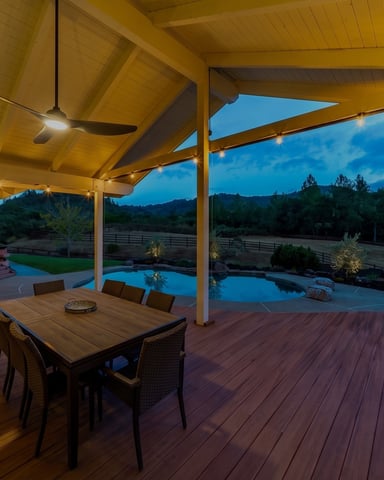 Covered deck with dining table and string lights overlooking a pool and hillside at dusk