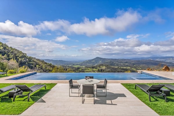 Infinity pool overlooking rolling hills and valleys, with patio seating and lounge chairs on manicured lawn