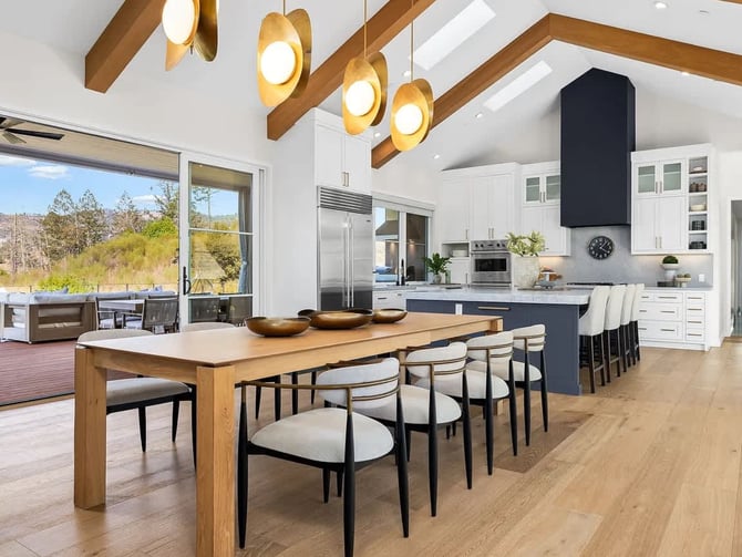Modern open-plan outdoor kitchen and lounge with blue cabinetry, built-in grill, dining benches, and seating area under vaulted ceiling with skylights